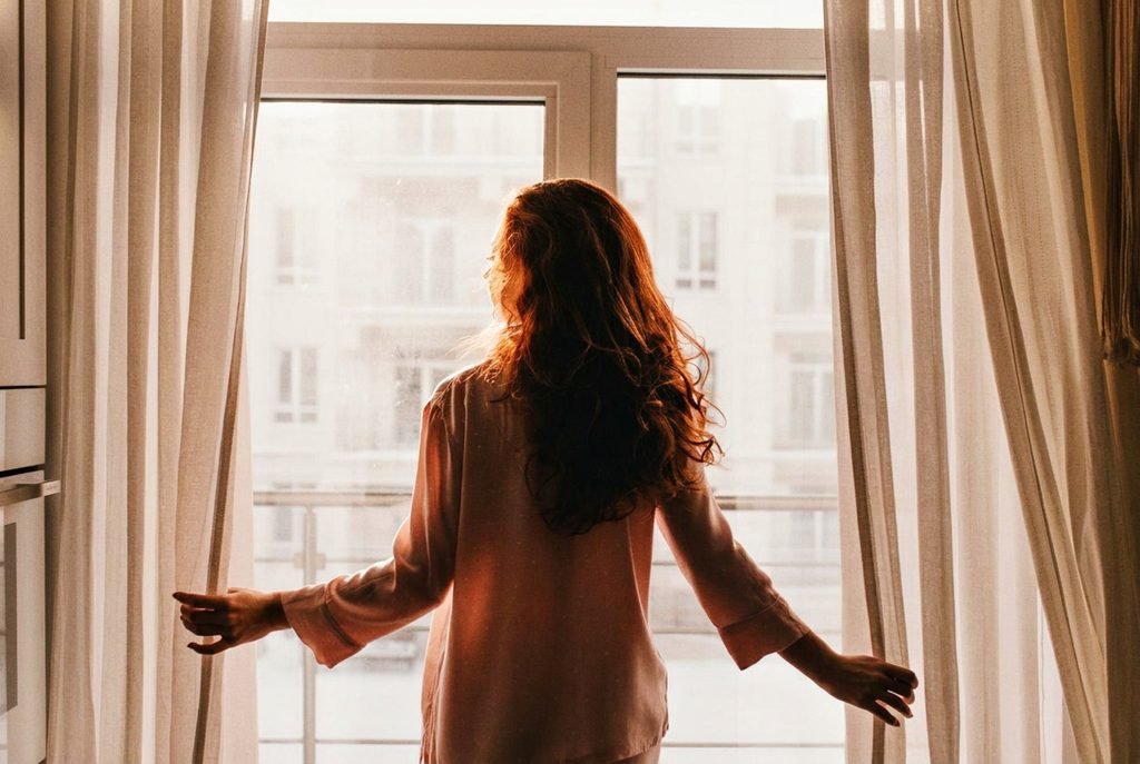 Woman opening the window curtain on a sunny Sunday morning.